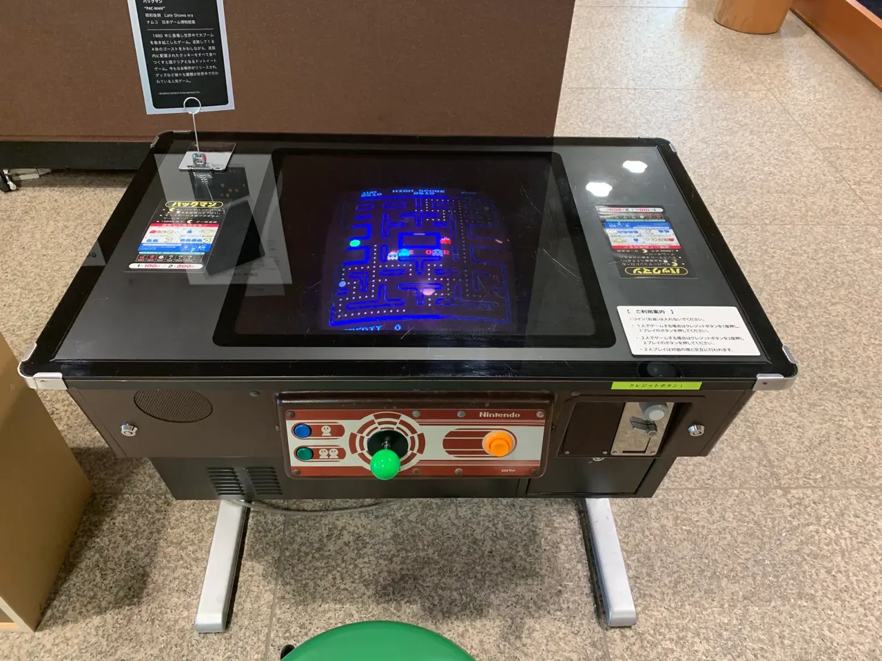 Müze ortamında duran renkli bir oyun kabini; yan panelinde Pac-Man figürleri ve hayaletler resmedilmiş.