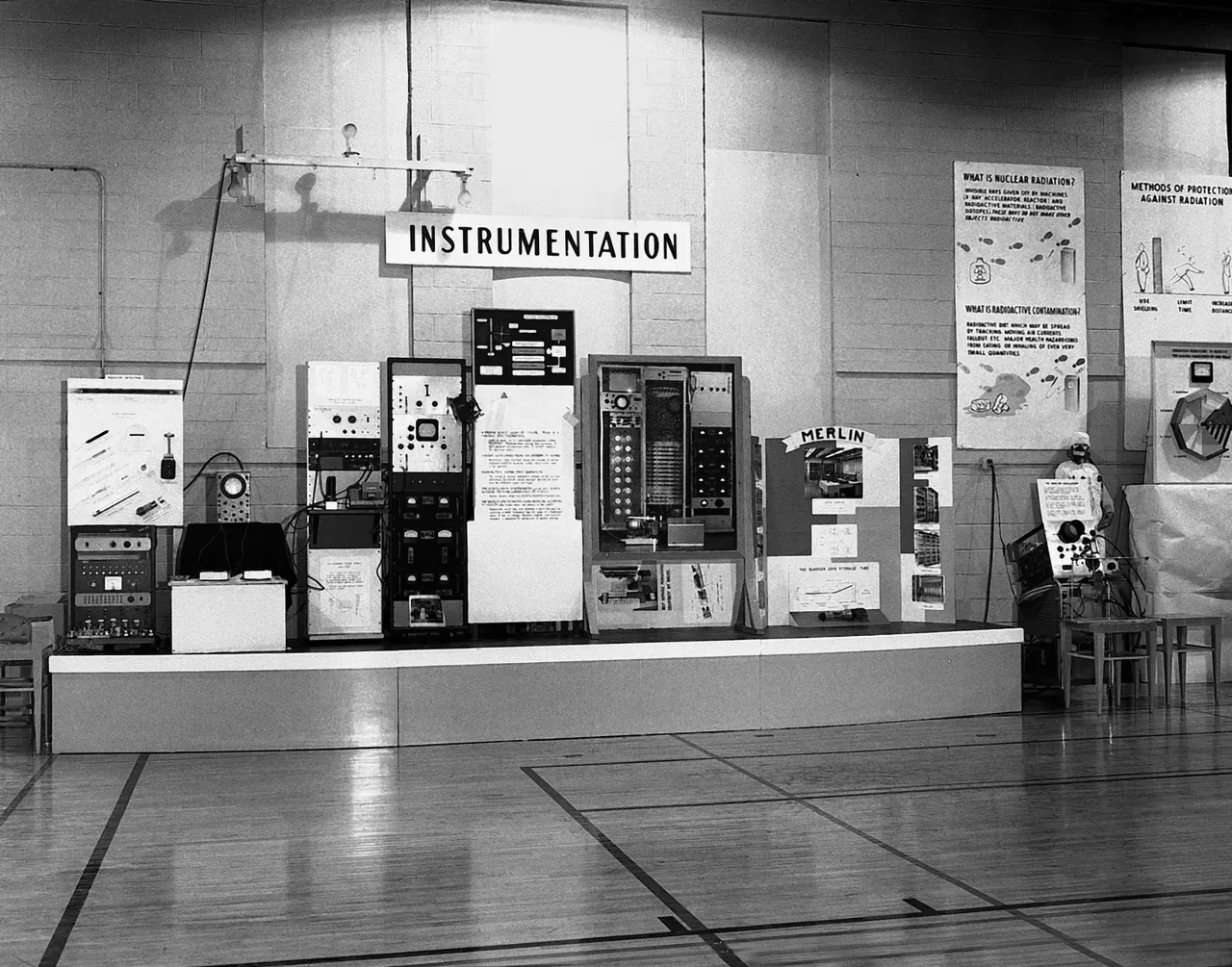 1950'ler siyah beyaz fotoğraf: bir spor salonunda kurulmuş sergi platformu; üzerinde sıra sıra laboratuvar cihazları, arkada “INSTRUMENTATION” yazan bir tabela ve radyasyon konulu bilgi afişleri.