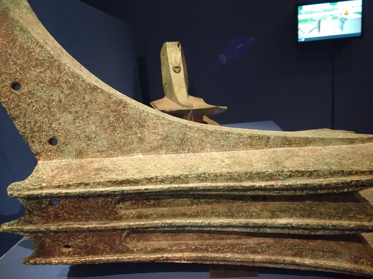 Cam vitrinde, yeşil patina kaplı tunç bir gemi mahmuzu; öne doğru sivrilen üç yatay bıçak, yan yüzeyinde kabartma bir yazıt.