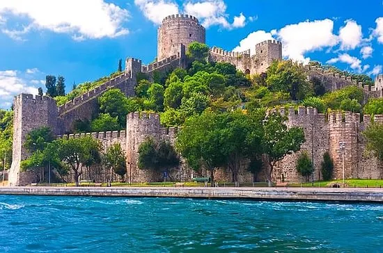 Boğaz kıyısında, yeşil tepe üzerinde kuleli taş kale; önünde mavi su.