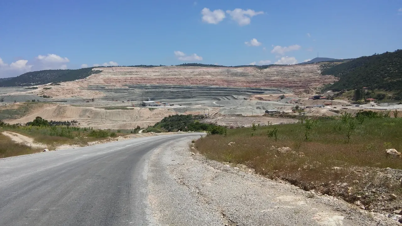 Bir tepenin içine oyulmuş geniş, basamaklı açık ocak; soluk pembe-beyaz-gri şeritli katmanlar teraslar hâlinde üst üste diziliyor, tabanda koyu pasa yığınları ve küçük iş makineleri, iki yanda çam kaplı sırtlar.