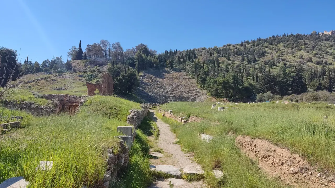 Yamaca oyulmuş yarım daire biçimli antik tiyatronun taş basamakları; arkada zeytinlikler ve ova.