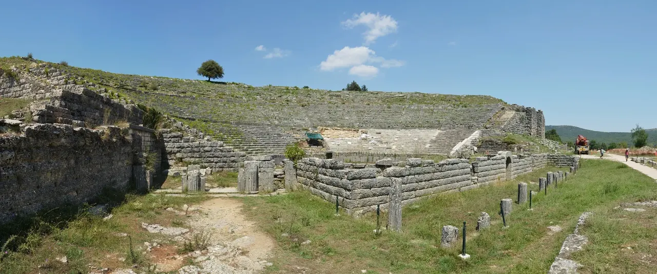 Dik yamaca kurulmuş çok basamaklı antik tiyatronun geniş açı görüntüsü; arkada dağlar ve yeşil vadi.