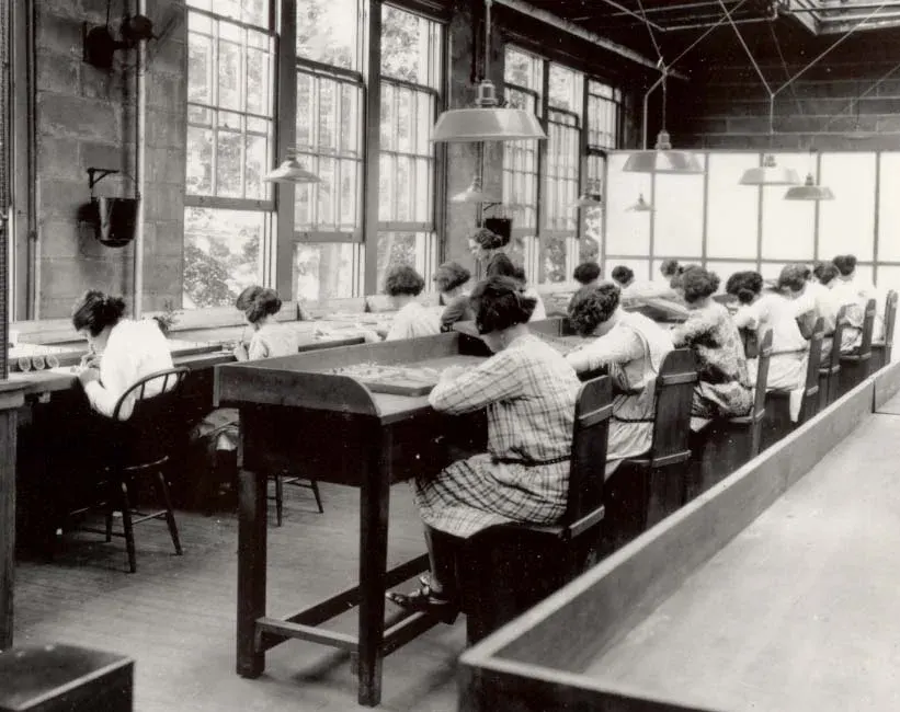 Siyah beyaz fabrika fotoğrafı: uzun masalarda yan yana oturmuş, önlerindeki küçük parçalar üzerinde çalışan genç kadınlar.
